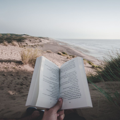 Grafika reklamująca książki na wakacje: osoba siedząca na plaży trzyma w dłoni otwartą książkę. 