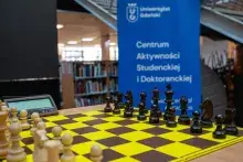 Na zdjęciu zbliżenie na szachownicę rozłożona na stole, w tle niebieski roollup Centrum Aktywności Studenckiej i Doktoranckiej fot. 
