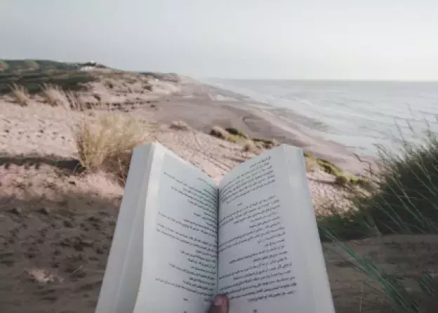 Grafika reklamująca książki na wakacje: osoba siedząca na plaży trzyma w dłoni otwartą książkę. 
